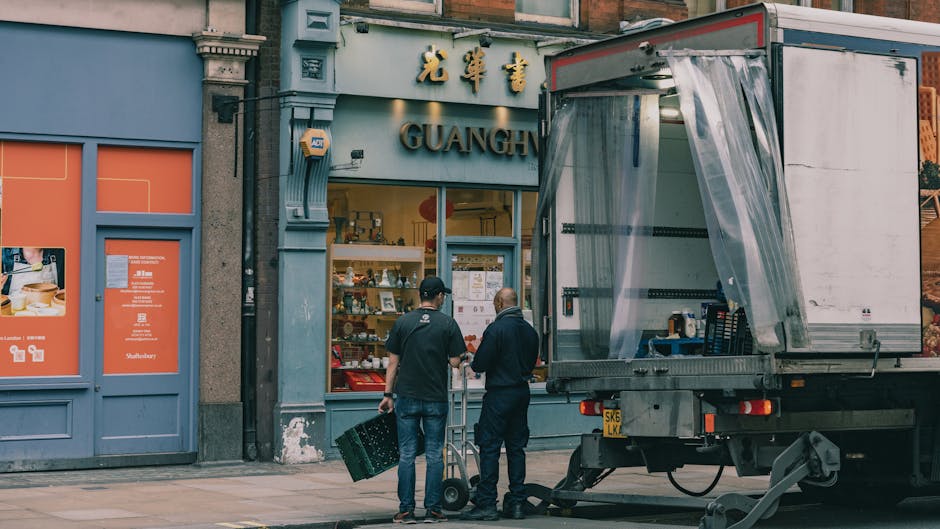Two removal men are preparing for a house relocation outside a shop on Frith Street near Carnaby Street, with a partially open moving truck parked on the pavement. One man is holding a black plastic c