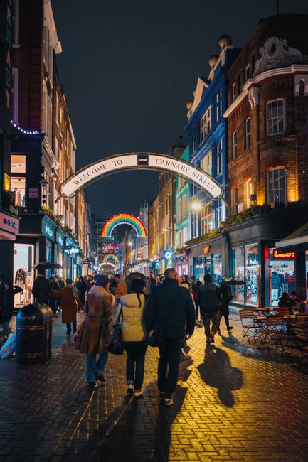 Two removal men are preparing for a house relocation outside a shop on Frith Street near Carnaby Street, with a partially open moving truck parked on the pavement. One man is holding a black plastic c